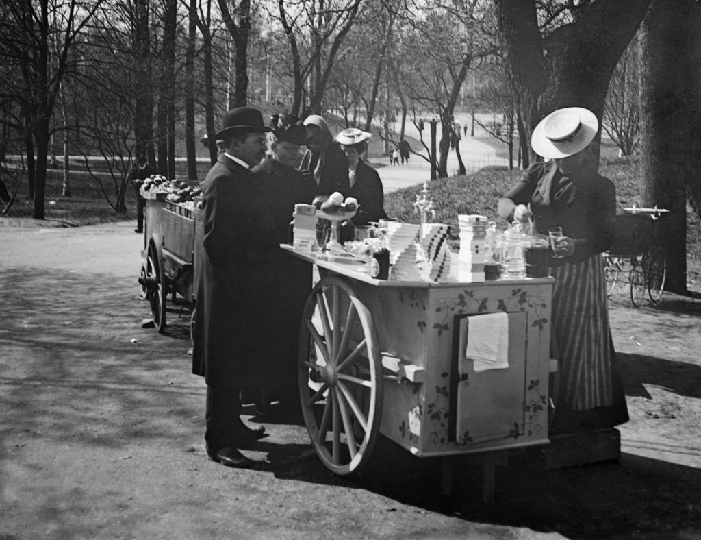 Helsingin muinaismuistolautakunta ja Signe Brander (keskellä) sunnuntairetkellä Kaisaniemessä virvokkeiden myyntikärryn luona. Kuva: Reinhold Hausen 1907