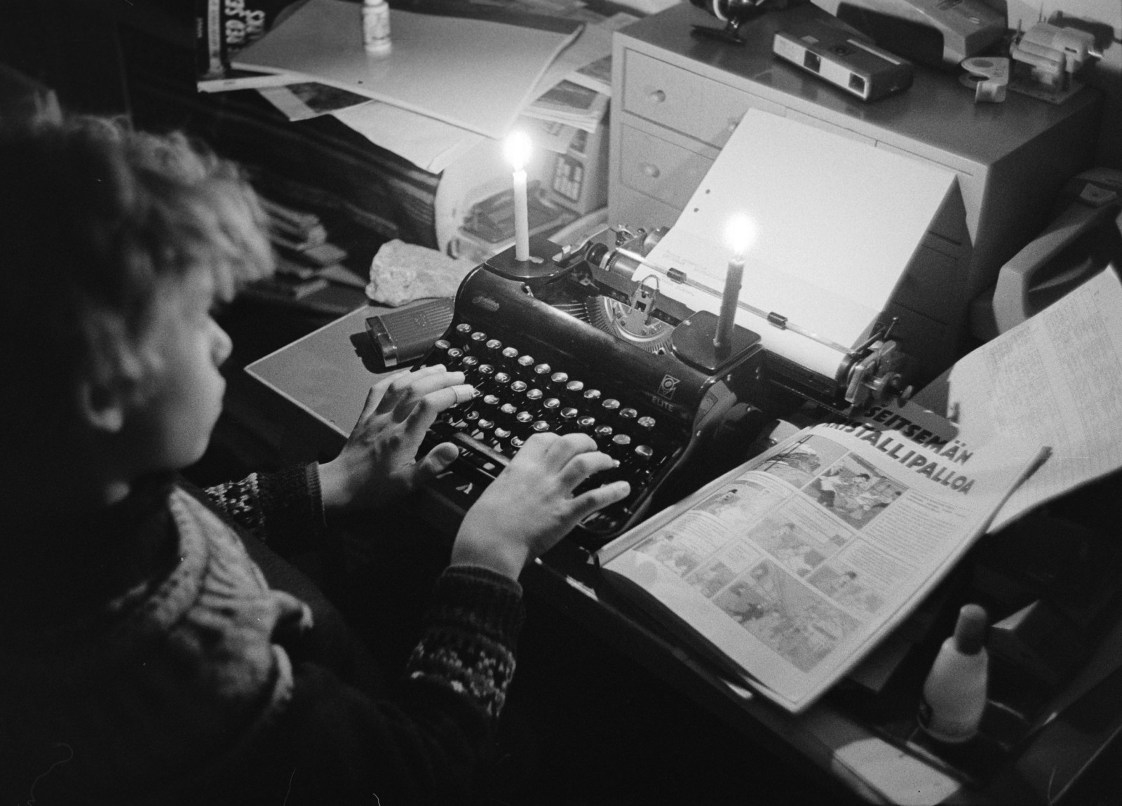 Poika kirjoittaa mekaanisella kirjoituskoneella kynttilänvalossa. Kuva: Helsingin kaupunginmuseo/Lauri Pietarinen, 1970-luku.